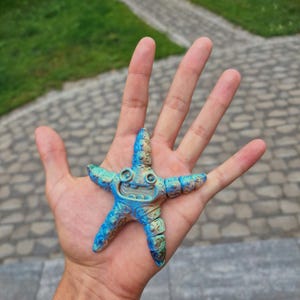 Puede incluir: Una pequeña figura de estrella de mar articulada con un degradado de colores azul y dorado. La criatura tiene una boca ancha y sonriente con dientes visibles, dos ojos redondos prominentes y una superficie texturizada con extremidades segmentadas.