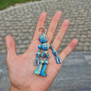 Puede incluir: Un pequeño llavero de robot articulado con un degradado de colores azul y beige. El robot tiene una cabeza cuadrada con dos ojos circulares, un torso rectangular y extremidades articuladas que terminan en manos y pies en forma de garra.