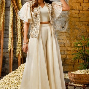 Puede incluir: Un conjunto de tres piezas color crema compuesto por un top corto sin mangas, pantalones anchos con una capa superpuesta transparente y una chaqueta de manga corta con intrincados bordados florales dorados y plateados y volantes.