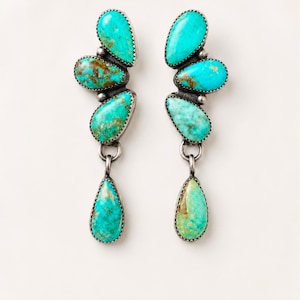 Può includere: Un paio di orecchini pendenti con diverse pietre di turchese a forma di goccia incastonate in argento. Ogni orecchino è composto da tre pietre sovrapposte collegate da maglie in argento, con una pietra a goccia che pende in fondo.