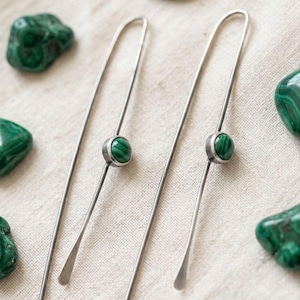 May include: A pair of long, slender silver-toned metal earrings featuring a single round, polished green malachite stone set in the center of each wire. The earrings have a minimalist, elongated U-shape design with a smooth, metallic finish.