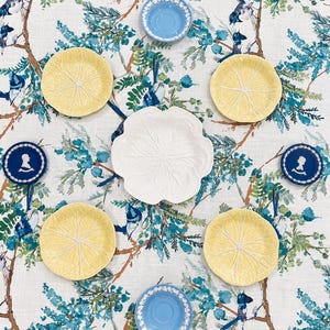 May include: A collection of ceramic tableware arranged on a white fabric with a blue and green bird and branch pattern. The set includes four yellow lemon-shaped plates, one white flower-shaped plate, two blue saucers, and two dark blue cameo plates.