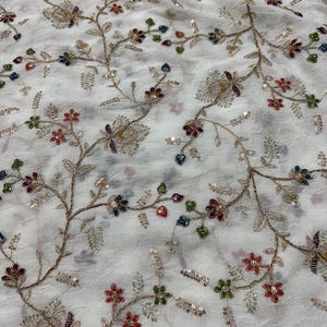 Könnte beinhalten: Ein weißer Stoff mit einem komplizierten Stickmuster aus gewundenen Zweigen mit kleinen roten, blauen und grünen Blumen. Das Design ist mit metallischem Goldfaden und kleinen, reflektierenden Pailletten akzentuiert, die über die Blumenmotive verteilt sind.