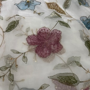 Puede incluir: Una tela blanca transparente con un intrincado patrón floral de flores bordadas en malva, azul y verde. El diseño incluye delicados detalles en hilo dorado, pequeñas lentejuelas transparentes y cuentas que acentúan los pétalos y las hojas.