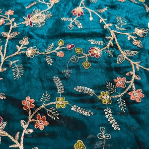 Op de afbeelding: Een diep teal-kleurige stof met ingewikkeld goudkleurig borduurwerk in een uitgestrekt bloemenrankpatroon. Het ontwerp bevat kleine bloemen in roze, paarse en groene tinten, geaccentueerd met kleine metalen lovertjes verspreid over de stengels.