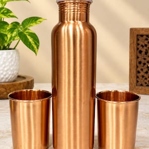 Pode incluir: Uma garrafa de água de cobre polido com tampa de rosca a condizer, acompanhada por dois copos de cobre a condizer. O conjunto apresenta um acabamento metálico liso com um corpo cilíndrico e um gargalo estreito, refletindo a luz na superfície.