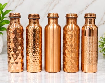 Puede incluir: Cinco botellas de agua cilíndricas de cobre dispuestas en fila. Cada botella presenta una textura exterior distinta: patrones de ondas verticales, acabado martillado, superficie lisa pulida, diseño hexagonal de panal y banda grabada.