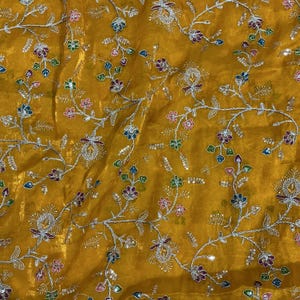 Könnte beinhalten: Ein senfgelber Stoff mit einem komplizierten Stickmuster aus silbernem Garn, das sich windende Ranken und kleine Blumen darstellt. Die Blumenmotive sind mit winzigen, bunten Pailletten in Rosa, Blau und Grün verziert, was eine strukturierte Oberfläche ergibt.