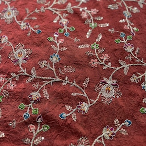 May include: A deep maroon fabric with a crinkled texture featuring intricate silver thread embroidery. The design consists of winding vines and floral motifs accented with small, colourful sequins in shades of blue, green, pink, and purple.