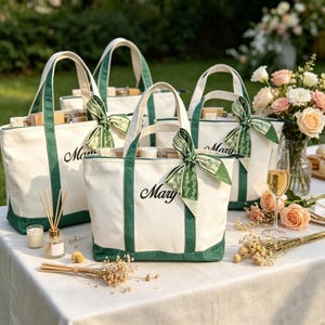 Può includere: Diverse borse tote in tela bianca con finiture e manici verdi sono disposte su un tavolo. Ogni borsa porta il nome "Mary" scritto in nero ed è adornata da un nastro verde. La scena è all'aperto, con fiori e un bicchiere di champagne.