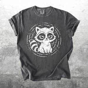 May include: A charcoal gray short-sleeve t-shirt featuring a white graphic print of a stylized raccoon with a cracked texture effect. The raccoon is centered within a circular frame composed of dashed lines and radiating crack patterns.
