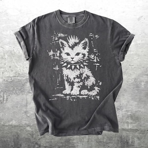 May include: A charcoal gray short-sleeve t-shirt featuring a white distressed graphic print of a kitten with a mohawk hairstyle and a spiked collar. The shirt has a crew neckline and rolled sleeves, showcasing a vintage-inspired washed finish.