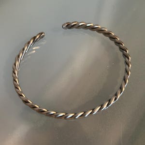 May include: A silver-toned metal cuff bracelet featuring a continuous twisted rope design. The bracelet is circular in shape with a small opening at the top, showcasing a polished metallic finish that reflects light along its spiraled surface.