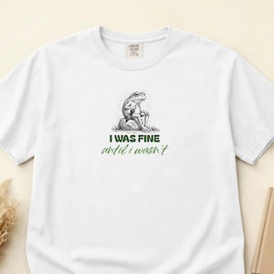 Op de afbeelding: Een wit t-shirt met korte mouwen voorzien van een zwart-witte schets van een kikker die op een rots zit en een paraplu vasthoudt. Onder de illustratie staat de tekst "I WAS FINE" in dikke groene letters en "until i wasn't" in groen cursief schrift.
