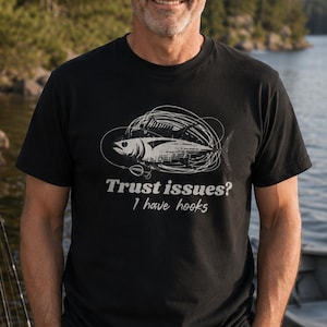 Può includere: Una t-shirt nera a maniche corte con una grafica bianca di un pesce circondato da lenze aggrovigliate e un amo. Il testo stampato in bianco recita: Trust issues? I have hooks. Il disegno è centrato sul petto del capo di abbigliamento.