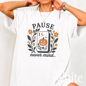 May include: A white t-shirt featuring a graphic print of a rectangular tarot card with a jack-o'-lantern, a crescent moon, and stars. The design is flanked by orange flowers and dark leaves, with the text PAUSE above and never mind... below.