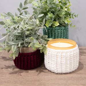 May include: Three potted plants with crocheted covers. The plants are in a variety of colors, including green, red, and white. The crocheted covers are a simple stitch pattern. The plants are sitting on a wooden surface.