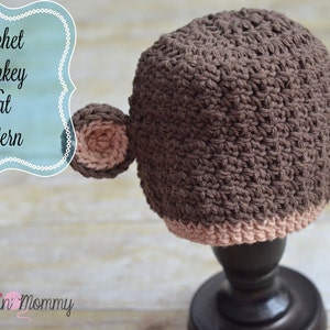 Może przedstawiać: Brązowa dzianinowa czapka z małpą z różowymi uszami. Czapka stoi na czarnym stojaku. Tekst "Crochet Monkey Hat Pattern" znajduje się na niebiesko-białym znaku.