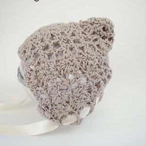 May include: A crocheted baby bonnet in a light beige color with a shell stitch pattern. The bonnet has a white satin ribbon tied at the bottom.