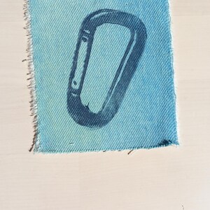 May include: A square-shaped light blue denim fabric patch featuring a dark blue screen-printed graphic of a carabiner. The patch has raw, frayed edges along the sides, showcasing the woven texture of the material and the distinct metal clip design.