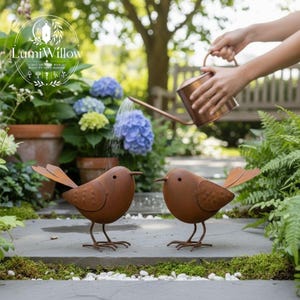 Könnte beinhalten: Zwei rostige braune Metallvogel-Skulpturen mit filigranen Beinen und Schwanzfedern stehen auf einem Steinweg. Die Vögel befinden sich in einem Garten mit Pflanzen und einer Person, die Blumen mit einer Kupfergießkanne gießt.