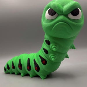 May include: A green caterpillar figurine with a large head, a grumpy facial expression, white eyes with black pupils, and a segmented body featuring black teardrop-shaped markings with small red centers along the sides and small protrusions.