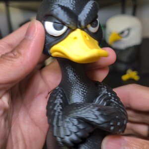 May include: A black duck figurine with a textured body, a bright yellow beak, and yellow webbed feet. The duck has a red crest on its head, white eyes with black pupils, and a stern facial expression with its wings crossed over its chest.