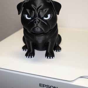 May include: A small, black, 3D-printed pug figurine with a grumpy facial expression, featuring white eyes and white-tipped paws. The dog is sitting in an upright position with its ears folded down, showcasing a textured surface finish.