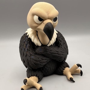 May include: A figurine of a vulture with a tan head, a white neck ruff, and black wings folded across its chest. The bird has a sharp black beak, narrow eyes with a stern expression, and tan legs with black talons, sitting in a seated position.