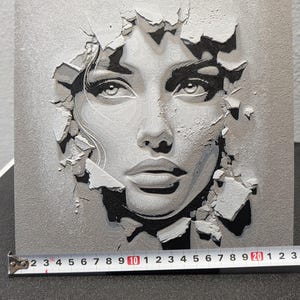 May include: A square wall art piece featuring a 3D relief portrait of a face emerging from a cracked surface. The artwork is rendered in shades of gray, white, and black. A metal measuring tape at the base shows a width of approximately 9.25 inches.