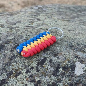 Puede incluir: Un llavero que presenta un dije rectangular de paracord trenzado con un patrón tricolor en azul, amarillo y rojo. El cordón está tejido en un diseño apretado y texturizado, unido a una argolla circular de metal plateado para las llaves.