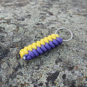 Op de afbeelding: Een sleutelhanger met een gevlochten paracord-ontwerp in felgeel en donkerpaars. Het koord is geweven in een dikke, getextureerde rechthoekige vorm en is aan één uiteinde bevestigd aan een zilverkleurige metalen ring voor veilige bevestiging.