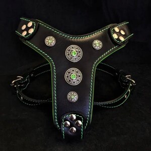 Op de afbeelding: Zwart leren hondentuig met groene stiksels en decoratieve zilverkleurige studs. Het tuig heeft een Y-vormig ontwerp, verstelbare riemen en metalen gespen. De studs hebben een groene edelsteen in het midden.
