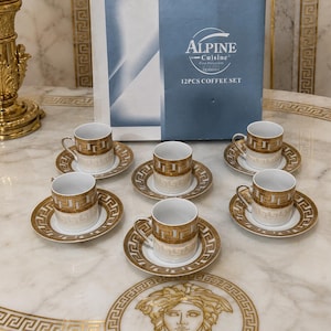 May include: A 12-piece coffee set by Alpine Cuisine featuring white porcelain cups and saucers with a gold Greek key pattern. The set includes six cups and six saucers, displayed in front of a blue and white box labeled Alpine Cuisine 12PCS COFFEE SET.