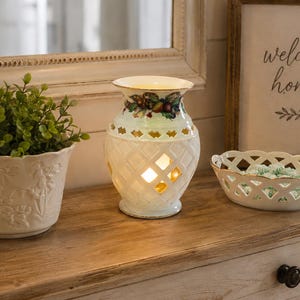 May include: A white ceramic wax warmer with a lattice pattern and a fruit motif rim, positioned between a white ceramic planter containing a green plant and a small white oval lattice bowl filled with light green decorative items.