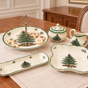May include: A collection of cream-colored ceramic tableware featuring a green Christmas tree motif with gifts, green trim, and holly accents. The set includes a two-tiered serving stand, a lidded sugar bowl, a creamer, a rectangular tray, and a bowl.