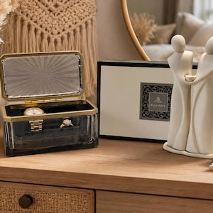 May include: A rectangular glass jewelry box with a gold-tone metal rim, containing a gold wristwatch and a ring, positioned next to a black and white rectangular box labeled PartyLite and a cream-colored abstract figurine of two figures holding hands.