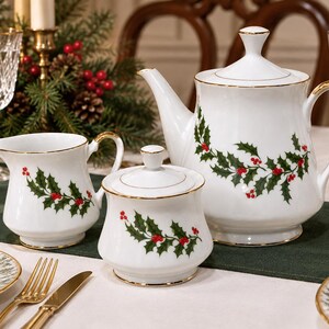 May include: A white porcelain tea set featuring a teapot, a creamer, and a lidded sugar bowl, each decorated with a green holly leaf and red berry pattern and finished with a thin gold rim along the edges of the pieces.