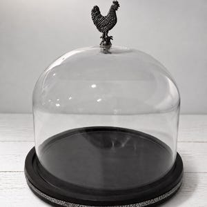 May include: A clear glass dome cloche with a dark, circular base featuring a textured silver-toned rim. A small, dark metal rooster figurine serves as the handle on top of the glass dome, providing a decorative accent to the functional food cover.