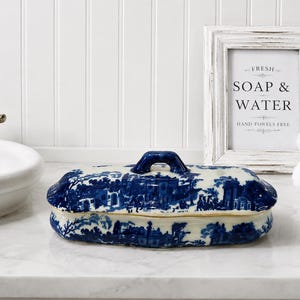May include: An oval ceramic soap dish with a lid, featuring a blue and white toile pattern depicting a landscape with buildings and trees. The dish has a small arched handle on the center of the lid and a scalloped edge along the base.