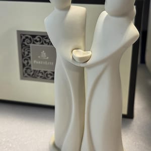 May include: A matte off-white ceramic figurine depicting two faceless figures standing together, holding a small heart-shaped bowl between them. The sculpture is positioned in front of a cream-colored box featuring the Partylite logo and text.