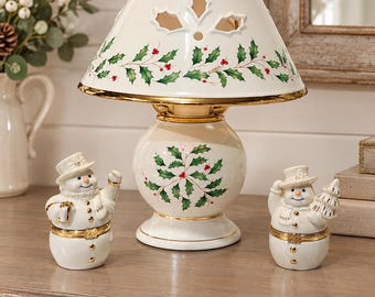 May include: A porcelain table lamp with a cream-colored base and shade, both decorated with green holly leaves and red berries. The lamp features gold trim. Two matching snowman-shaped porcelain trinket boxes stand on either side of the lamp.