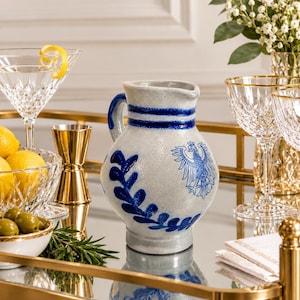 May include: A light gray ceramic pitcher with a blue handle and blue decorative bands around the neck. The body features a blue eagle emblem and a blue leaf pattern. The pitcher has a rounded shape and a small spout for pouring.