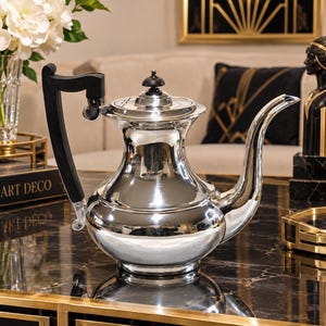 May include: A polished silver teapot featuring a rounded body, a long curved spout, and a black handle with a matching black finial on the lid. The metallic surface is highly reflective, showing a smooth, curved silhouette and a wide base.