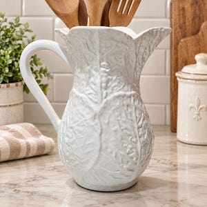 May include: A white ceramic pitcher featuring a textured, embossed leaf pattern across its exterior surface. The pitcher has a curved handle on the left side and a wide, scalloped rim. Several wooden kitchen utensils are placed inside the pitcher.