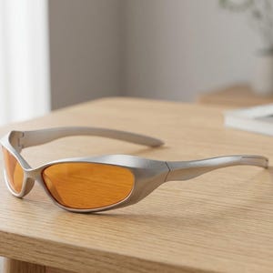 May include: Silver-framed sunglasses with orange lenses rest on a wooden surface. The sunglasses have a sleek, modern design with curved arms and a streamlined shape. The lenses are a vibrant orange color.