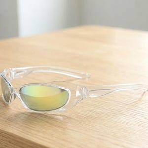 Puede incluir: Gafas de sol de plástico transparente con montura curva y lentes tintadas de amarillo. Las gafas de sol están sobre una superficie de madera clara. Las lentes reflejan un tono dorado.