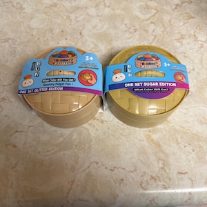 May include: Two round, tan-coloured plastic containers shaped like bamboo steamers, each featuring a blue paper band labelled Bun, One Set Glitter Edition, and One Set Sugar Edition. The packaging includes illustrations of colourful buns and text asking What Colour Will You Get? Squishy Fidget Fun.