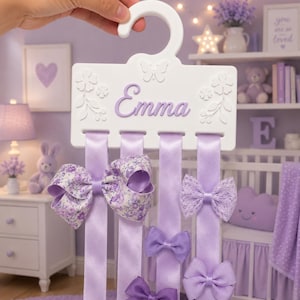 Pode incluir: Um organizador de laços de cabelo em madeira branca com gancho, apresentando o nome Emma em letra cursiva roxa. A placa é decorada com flores em relevo e uma borboleta. Quatro longas fitas de cetim roxo claro pendem com vários laços.