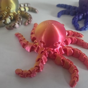 May include: Three 3D-printed octopus toys in various colours. The central octopus is a red-orange gradient, with gold and purple octopuses in the background. The toys have articulated tentacles and a detailed design.